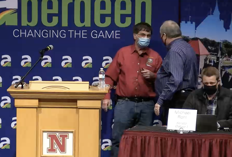 Sen. Al Novstrup, in red shirt, masked before start of Aberdeen crackerbarrel, speaking with fellow masked legislators Carl Perry (standing) and Michael Rohl (seated). Screen cap from Dakota Broadcasting video, 2021.01.30.