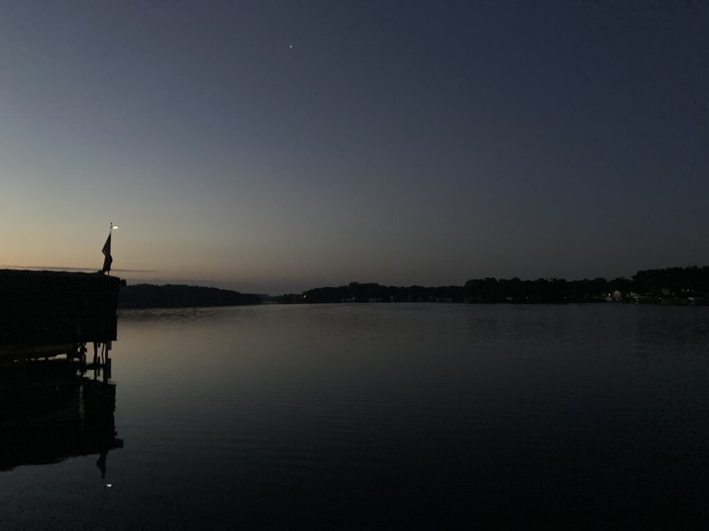 Dock with lighted flag, toward the southeast on Lake Minnewashta. The house next to the city campground stood empty, a summer home, it seems, larger than my last two houses combined. The Arnolds Park cemetery lies to the west, so the campground serves as a buffer between the summer house and a zombie attack, a free-fire zone where the seasonal residents can get clear shots at the heads of the walking dead. Maybe that thought is what kept me up much of the night.
